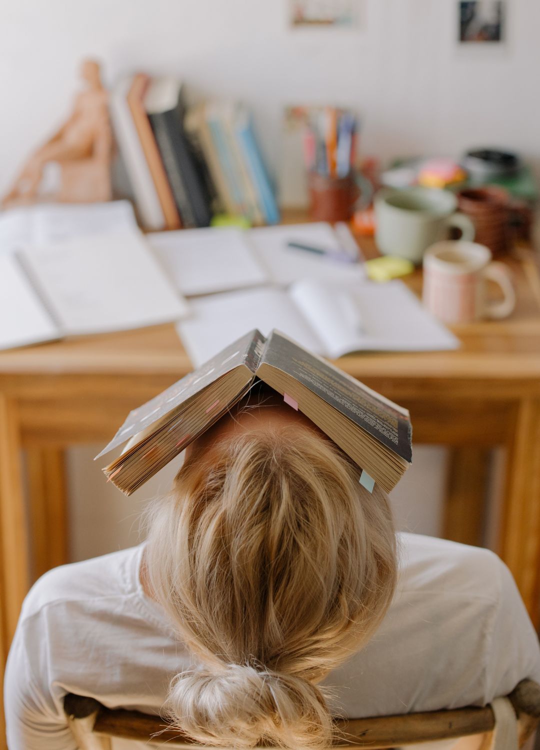 Vrouw zit voorovergebogen met een boek op haar hoofd bij stress door studie of werkdruk.
