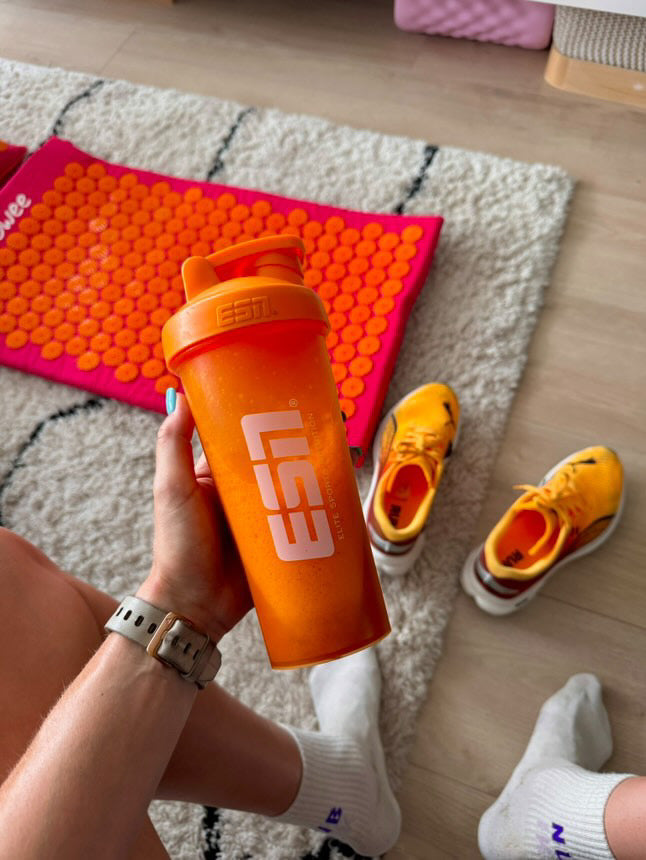 Sporter met shakebeker naast sportschoenen en een Flowee spijkermat voor herstel na het sporten.