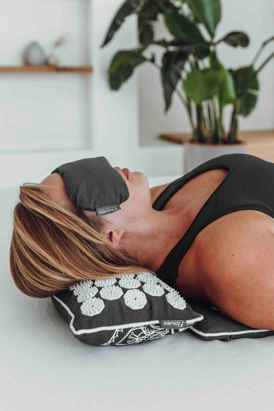 Vrouw ligt op spijkermat met kussen in de kleur grijs met wit. Met op haar ogen een oogkussen in de kleur grijs.
