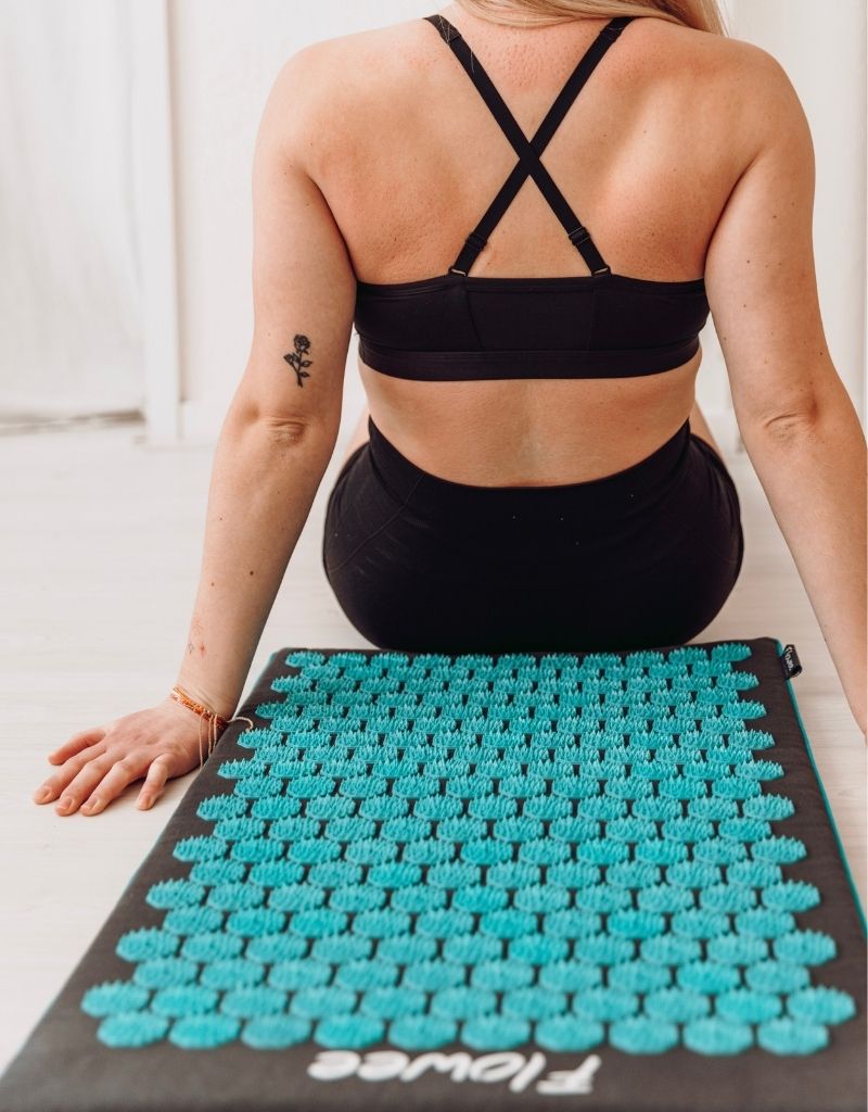 Vrouw zit naast een Flowee spijkermat met zeegroene spikes voor ontspanning bij rugklachten.