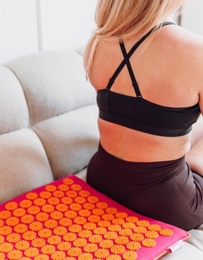 Vrouw zit naast een Flowee spijkermat met oranje spikes voor ontspanning bij rugpijn.