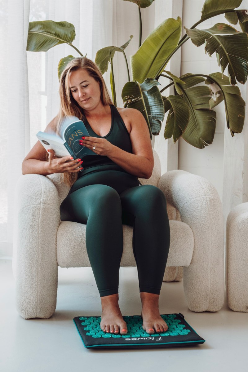 Een vrouw in groene sportkleding zit op een stoel een boek te lezen en tegelijkertijd met het voeten op een grijs spijkermatje met zeegroene spikes.