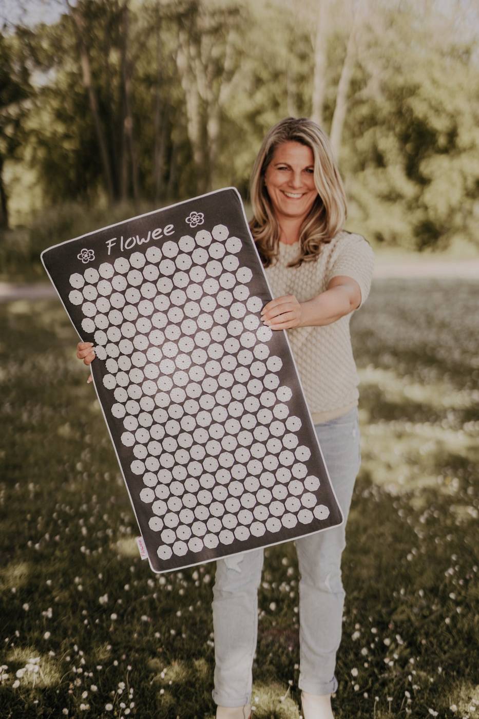 Vrouw staat lachend met een zwarte spijkermat met witte spijkers van Flowee in haar handen op een grasveld.