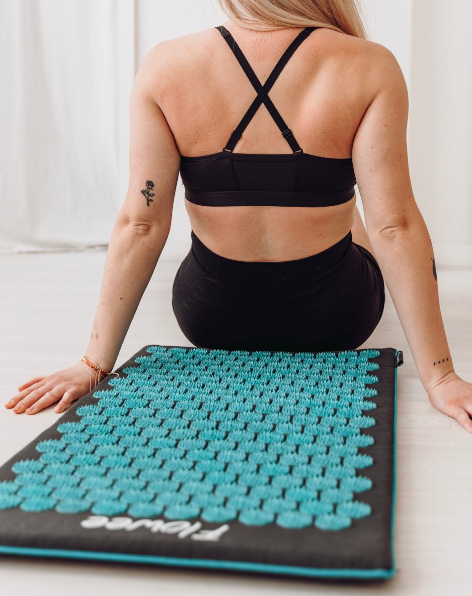 Een vrouw zit voor een grijze acupunctuur mat met zeegroene spikes, waarbij het resultaat in haar rug te zien is.
