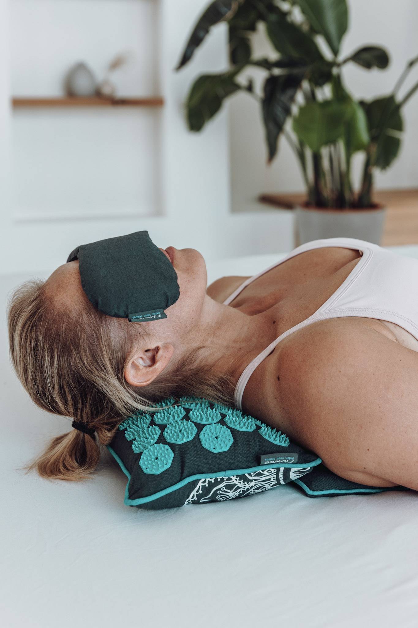 Een vrouw ligt op een grijze spijkermat en kussen met zeegroene spikes op een bed met een oogkussen op