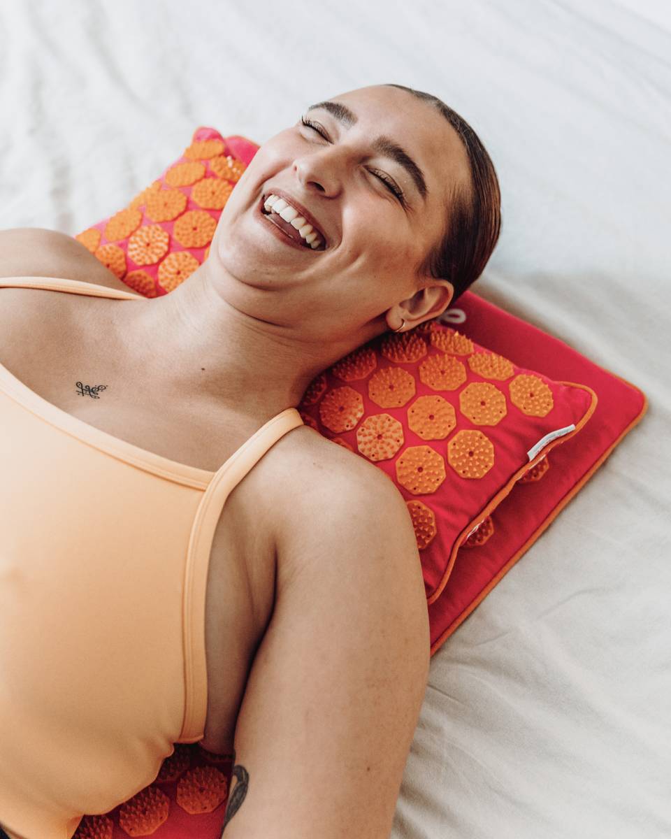 Een lachende vrouw in oranje sportkleding ligt op een spijkermat eco set fuchsia met oranje op een wit dekbed