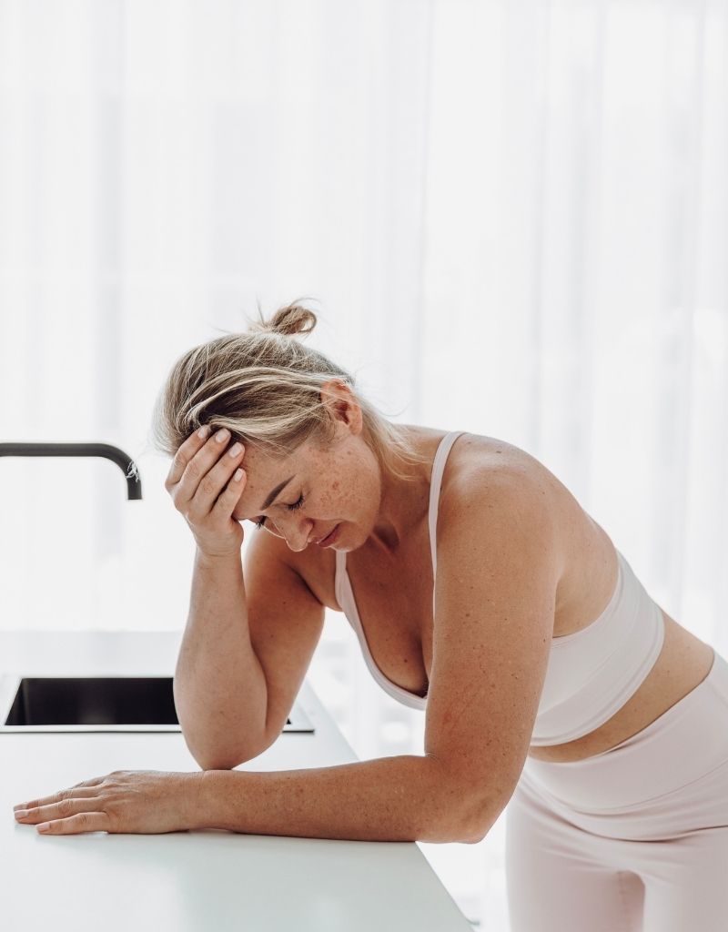 Vrouw leunt vermoeid op het aanrecht en houdt haar hoofd vast bij overgangsklachten.