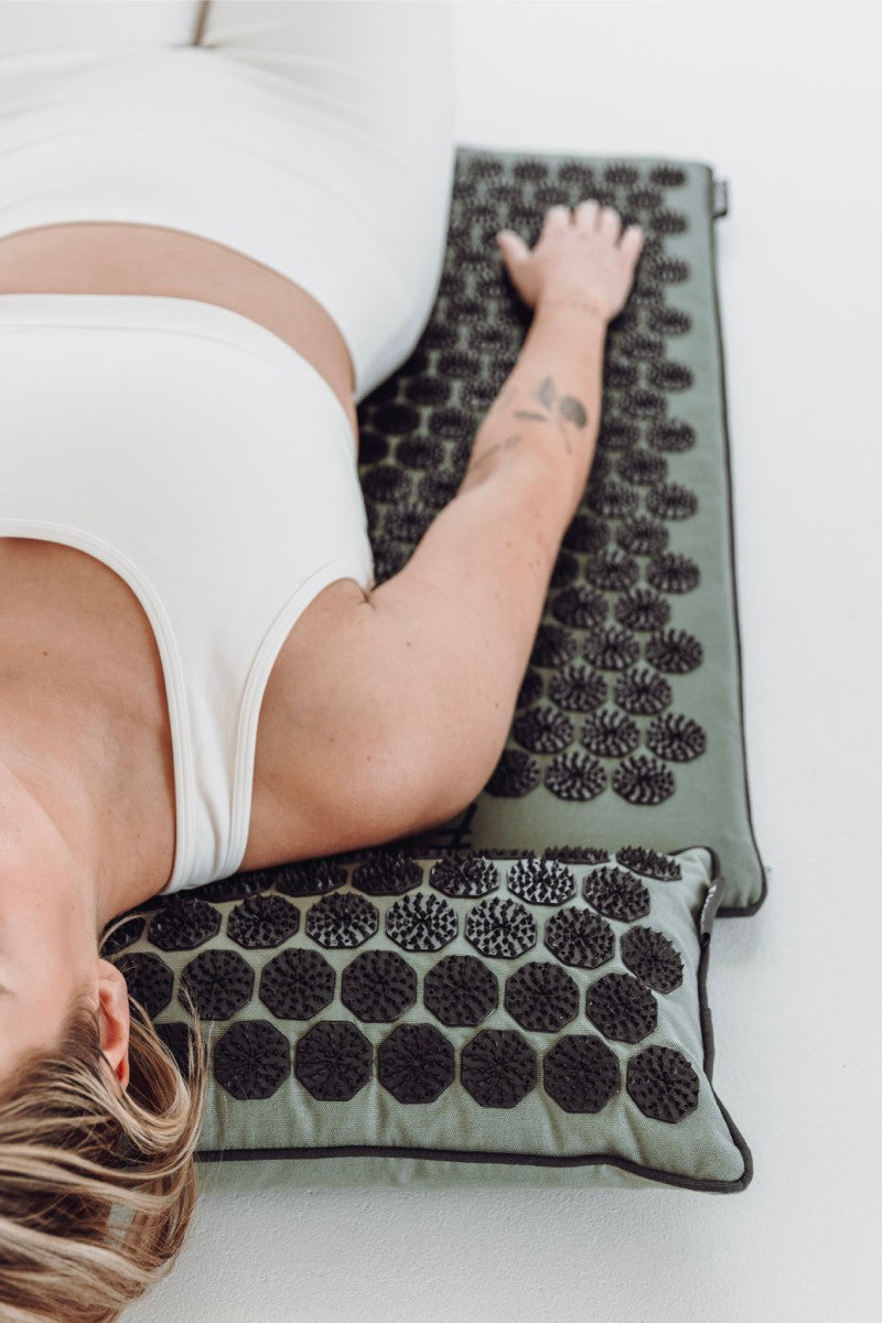 Een vrouw in een wit sporttenue ligt op een groene shakti mat met grijze spikes op een witte vloer.