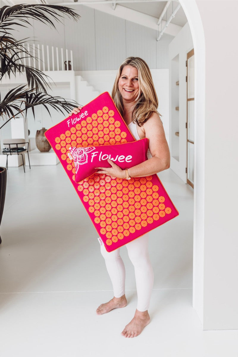 Een vrouw staat in een grote kamer in een wit sporttenue met een fuchsia shakti mat en shakti kussen met oranje spikes in haar handen.