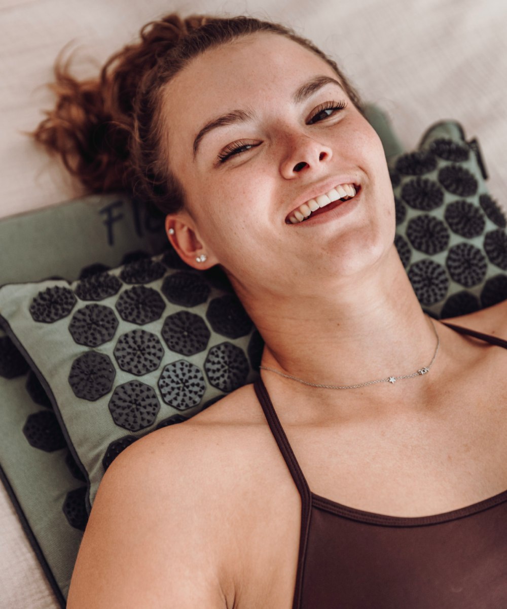 Een vrouw ligt lachend op een groene shakti mat en shakti hoofdkussen met grijze spikes.