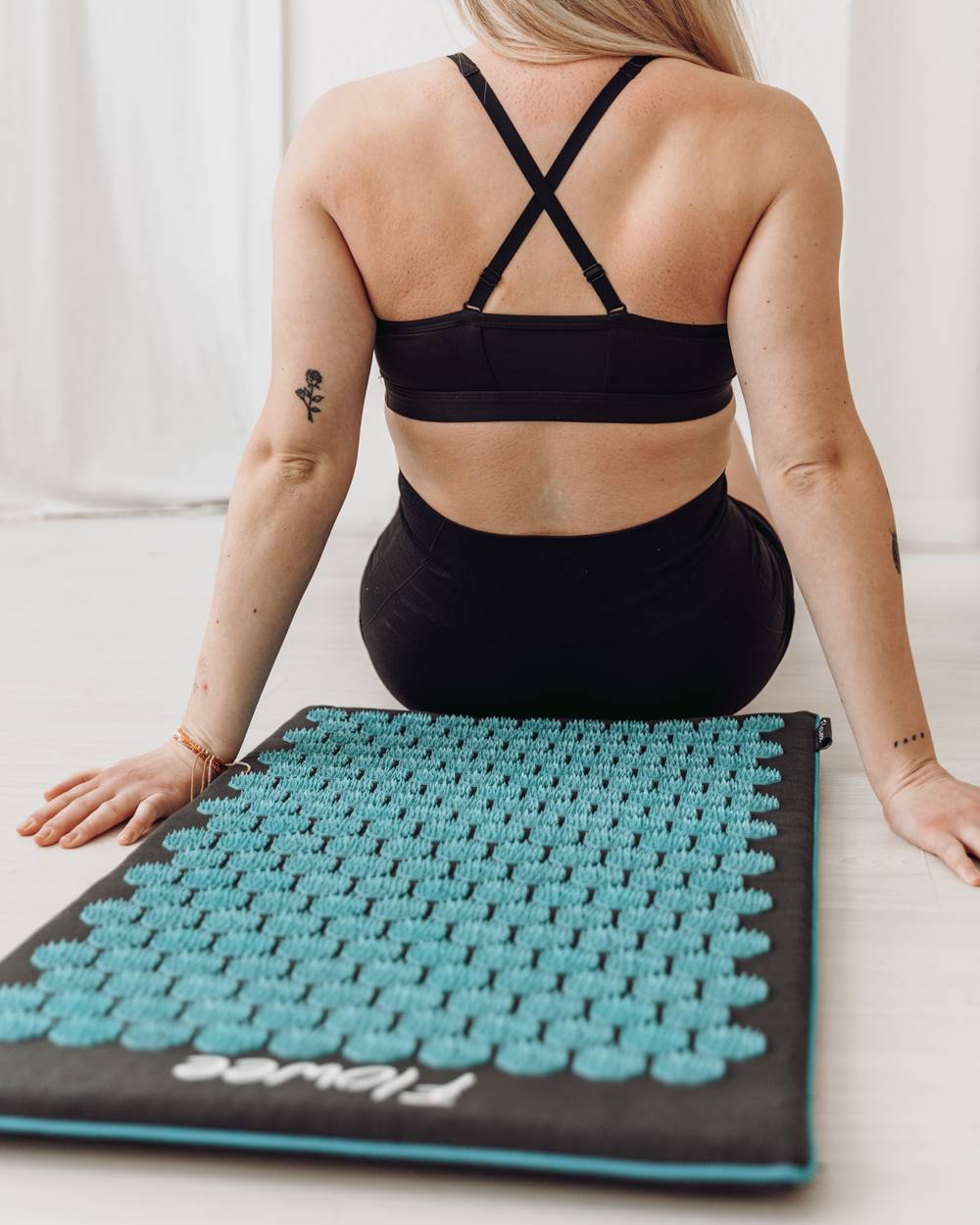 Een vrouw met zwarte kleding aan zit voor een grijze spijkermat met zeegroene spikes van Flowee, met het resultaat zichtbaar op de rug.