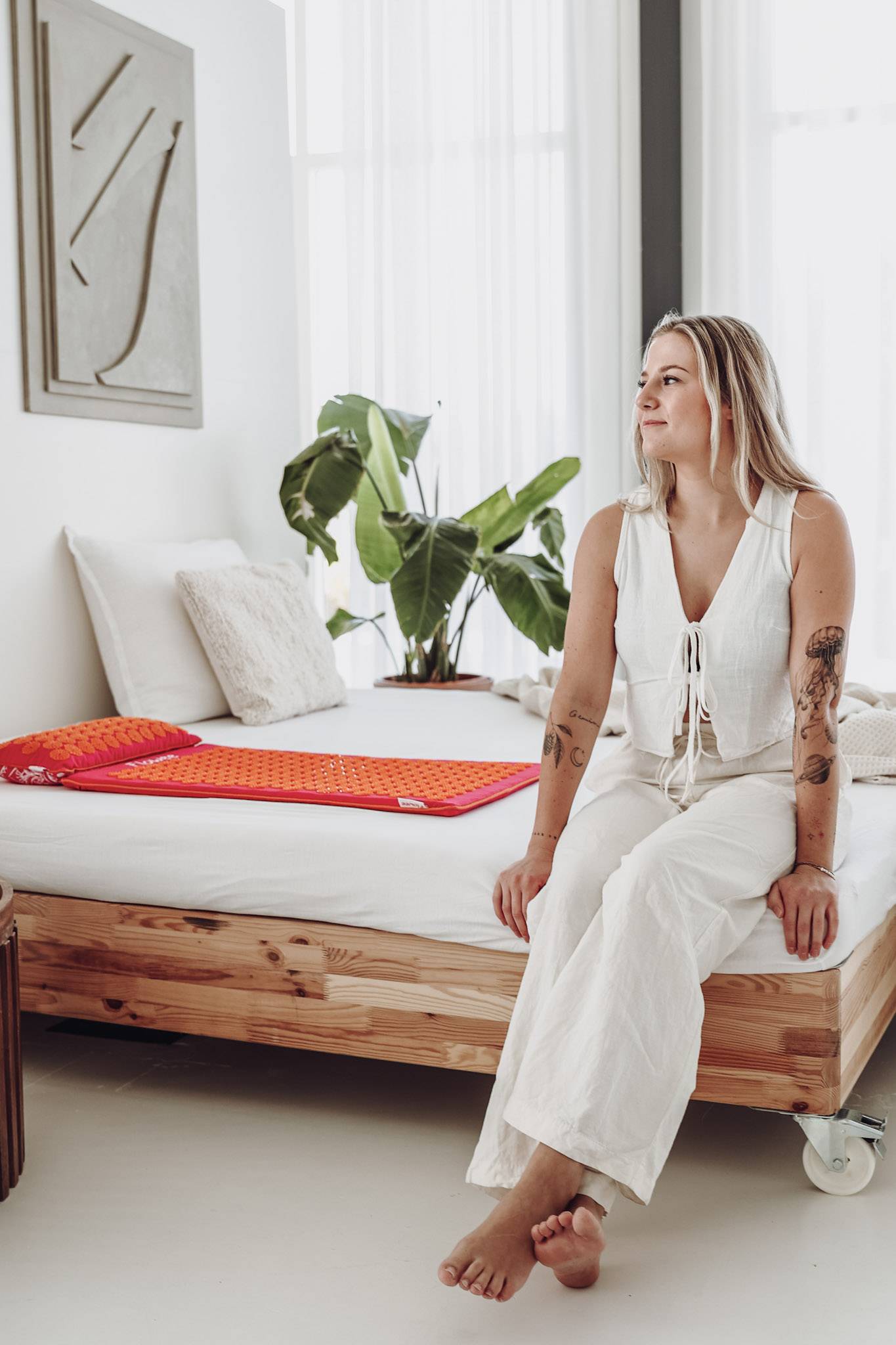Een vrouw in witte kleding zit op bed waarnaast een fuchsia spijkermat met oranje spikes ligt