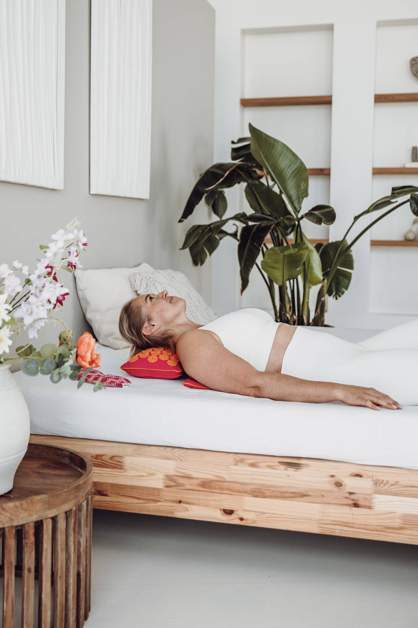 Een vrouw in witte sportkleding ligt op een spijkermat op het bed met eromheen planten en bloemen
