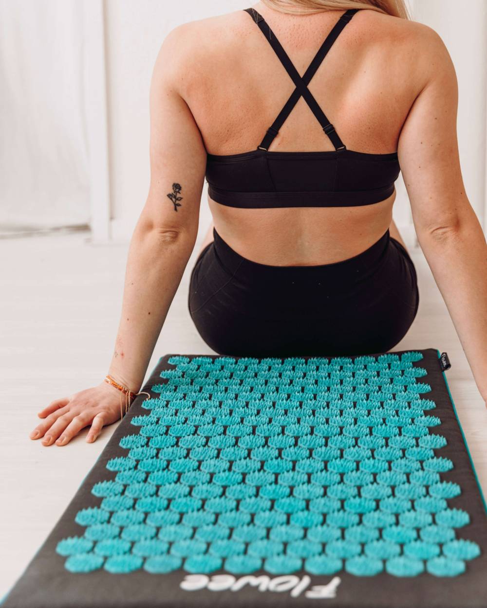 Een vrouw zit rechtop voor een zwarte Flowee spijkermat met zeegroene spijkers waarbij het resultaat in haar rug staat.