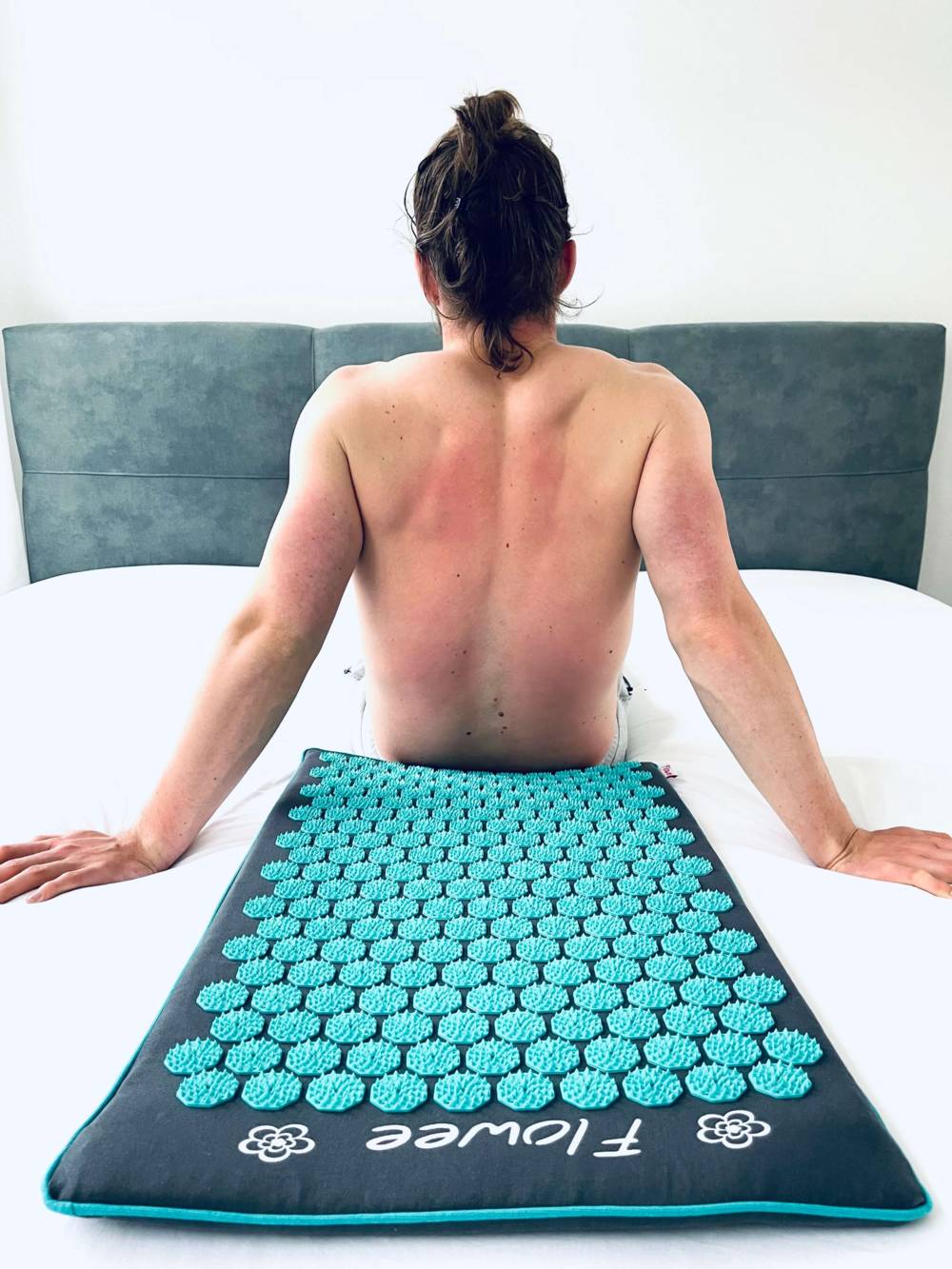 Een man zit rechtop op een bed voor een grijze spijkermat met zeegroene spijkers van Flowee met het resultaat van de spijkers in de rug.