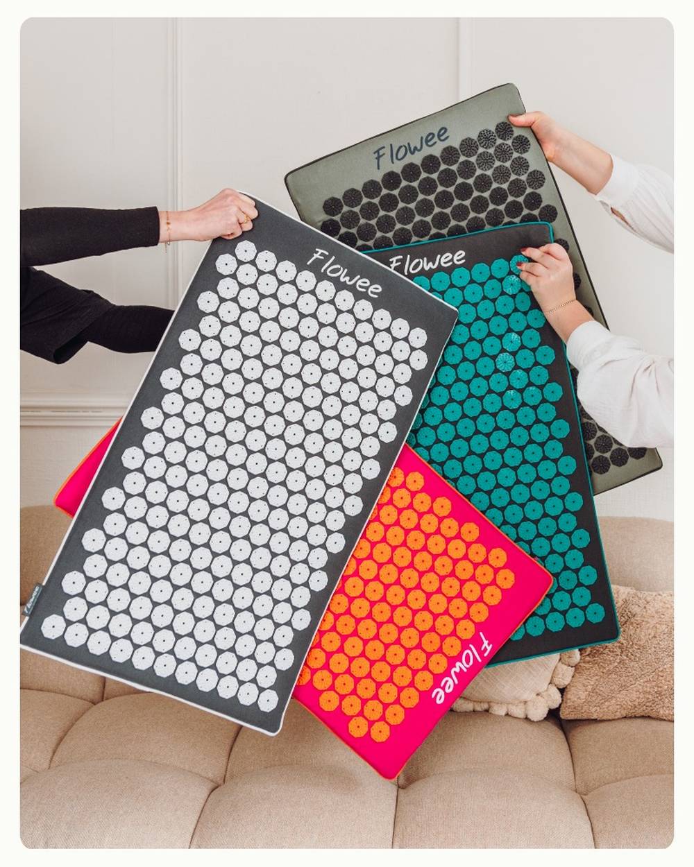 Twee vrouwen waarvan alleen de armen in beeld zijn houden 4 spijkermatten van Flowee vast in verschillende kleuren boven een bank.