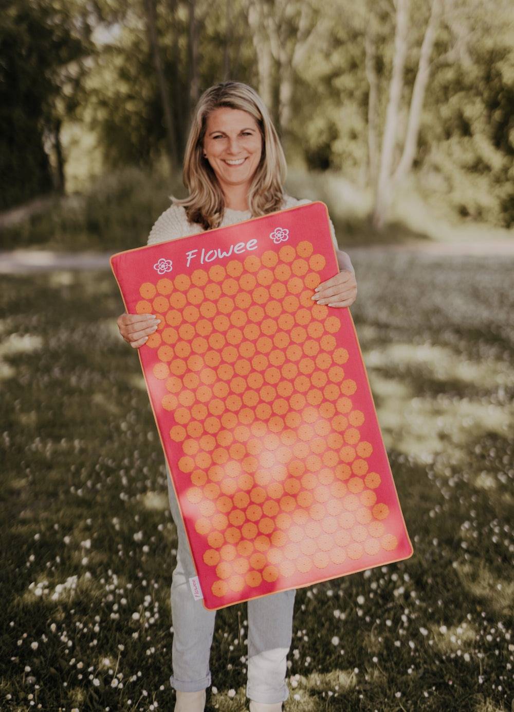 Lachende vrouw met een fuchsia oranje spijkermat van Flowee in haar handen staand in een grasveld.