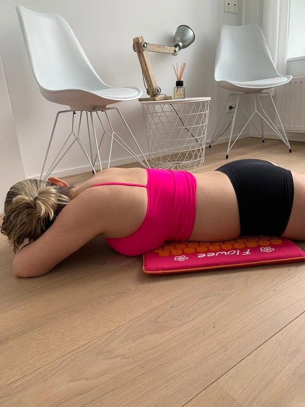 Een vrouw in sportkleding ligt met haar buik op een fuchsia spijkermat van Flowee op een harde houten vloer.