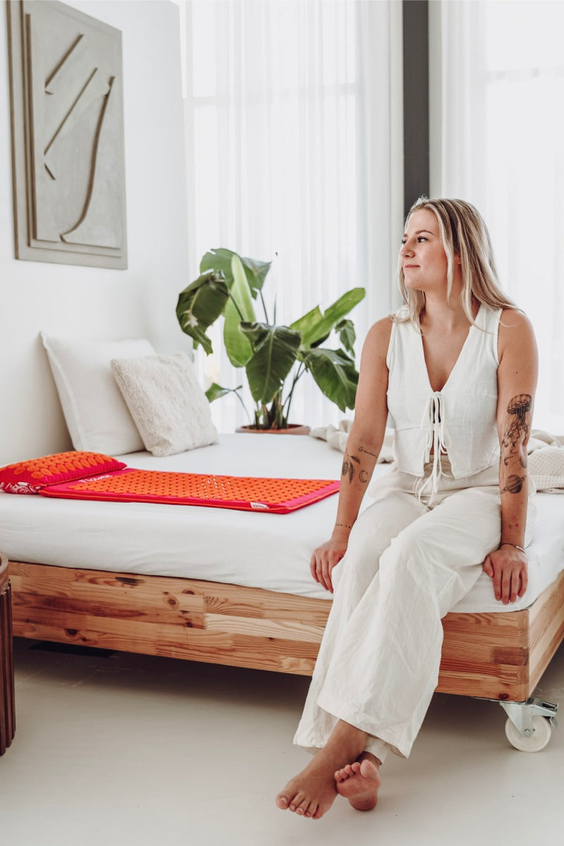 Een vrouw zit in een kamer op een bed waar een fuchsia shakti mat met oranje spikes op ligt.